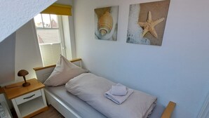 A small, single-bed room with a wooden nightstand, a white drawer, and a wall-mounted shelf. There are two wall art pieces depicting shells and a starfish.