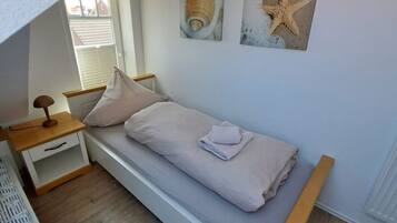 Ein kleines Zimmer mit einem Einzelbett, einem Nachttisch aus Holz, einer weißen Schublade und einem an der Wand befestigten Regal. Zwei Wandbilder zeigen Muscheln und einen Seestern.