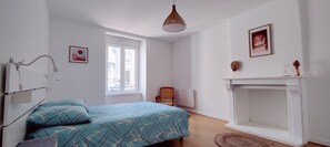 Un dormitorio con cama, ventana, radiador, silla, chimenea y lámpara colgante.
