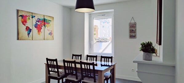 Un comedor con una mesa y sillas de madera, un cuadro con un mapa del mundo en la pared, una ventana con vista a edificios y una planta pequeña en un estante.