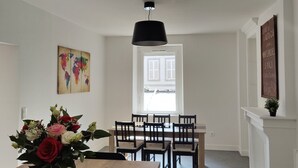 En matplats med ett träbord, stolar, en vas med blommor, en hängande lampa och ett fönster med utsikt över byggnader.