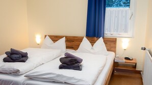 En dobbeltseng med hvide sengetøj, to puder og lagt sammen håndklæder. Der er et træhovedgærde, et natbord med en lampe, og et vindue med blå gardiner.