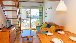 A cozy room with a wooden desk, a dining table set with plates and glasses, a sofa with cushions, a TV on a wooden cabinet, and a balcony with a view.