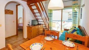 A dining area with a wooden table set for two, a television on a wooden cabinet, and a view of the outside balcony.