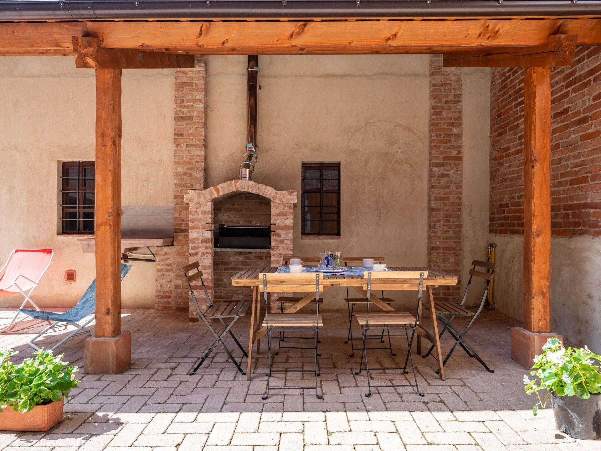 Eine Terrasse im Freien mit einem Backofen aus Ziegeln, einem Holztisch, Stühlen und Blumentöpfen.