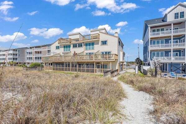 A beachfront hotel with multiple balconies, a sandy walkway, and a clear sky.