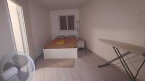Een kleine, smalle kamer met een bed, een ventilator, een bureau en een raam met luiken.