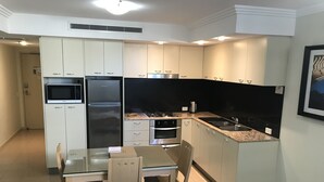 Ett kompakt kök med vita skåp, en mikrovågsugn, ett kylskåp, en spis och en diskho. Det finns ett matbord med stolar och en inramad tavla på väggen.
