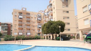 A swimming pool with steps leading into it, surrounded by apartment buildings with balconies. - 035 - Gaviotas 002 - comfortHOLIDAYS (Santa Pola)