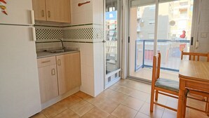 A compact kitchen with a refrigerator, sink, and wooden cabinets. A glass door leads to a balcony with a view of the city. - 035 - Gaviotas 002 - comfortHOLIDAYS (Santa Pola)
