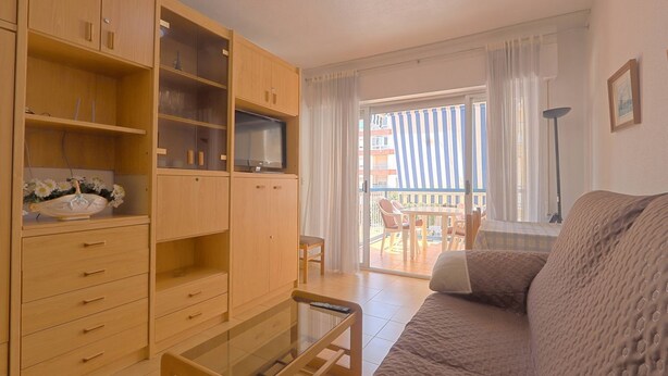 A living room with wooden cabinets, a glass coffee table, a sofa, and a balcony with a view of buildings. - 035 - Gaviotas 002 - comfortHOLIDAYS (Santa Pola)