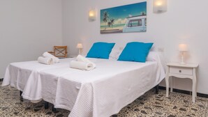 Ein Doppelbett mit weißer Bettwäsche und blauen Kissen, ein kleiner Nachttisch mit einer Lampe, ein Gemälde mit Strandmotiv und ein gefliester Boden.