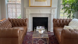 A living room with two brown leather sofas, a glass coffee table, a fireplace, and a framed painting above it.