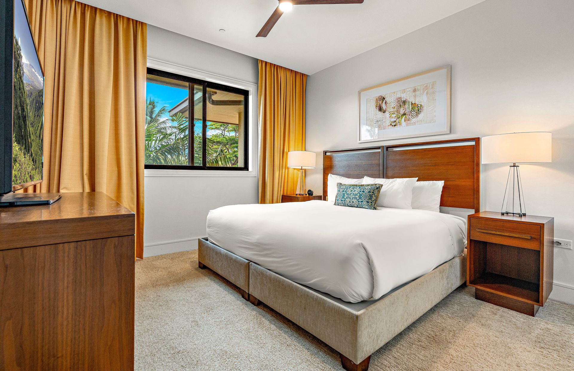 Une chambre d’hôtel avec un grand lit, un bureau, une table de chevet, un ventilateur de plafond, une fenêtre avec des rideaux et un tableau encadré au mur.