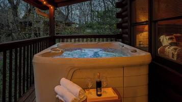 A hot tub with a bottle of wine and glasses on a wooden table, surrounded by a wooden deck with a railing.