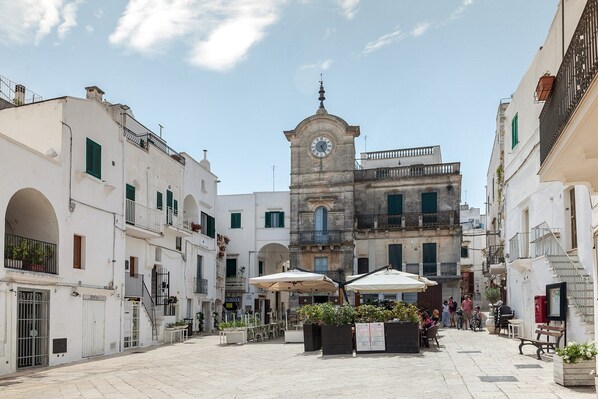 A square with a central clock tower, outdoor seating, and white buildings with green shutters. - Beautiful Garden Apartment in Cisternino (Cisternino)