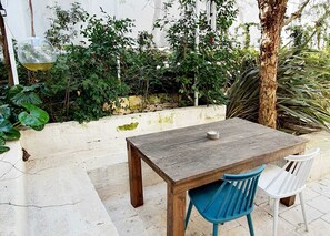 A wooden table with two chairs, one blue and one white, in an outdoor patio area with a white wall and greenery in the background. - Beautiful Garden Apartment in Cisternino (Cisternino)