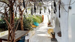 Un espacio de estar al aire libre con techo, que incluye una mesa y sillas de madera, rodeado de plantas en macetas y adornos colgantes.