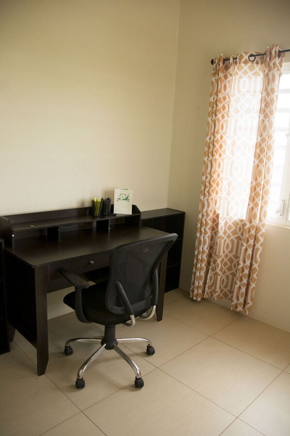 A dark wooden desk with a black office chair, a small shelf with pens and a notepad, and a window with patterned curtains. - Spectacular home -5 mins drive to beach, modern, comfortable, clean and spacious (Cul De Sac)