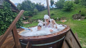 Un bain à remous en bois rustique, rempli d’eau et de mousse, entouré de verdure et doté d’une terrasse en bois.