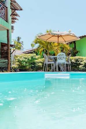 A pool area with a table and chairs under a canopy, surrounded by greenery and a building. - Hana Mana Flats (São Miguel dos Milagres)