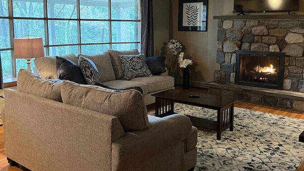 A cozy living room with a stone fireplace, a sofa with patterned cushions, and a large window offering a view of the outdoors. - 8 bed Lodge Near Camelback/Outlets-HOT TUB (Tannersville)