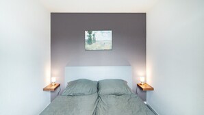 Una cama con un edredón gris, dos mesitas de noche con lámparas, un cuadro pequeño en la pared y un techo blanco liso.