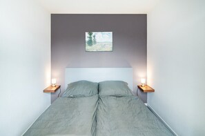 A bed with a grey blanket, two bedside tables with lamps, a small painting on the wall, and a plain white ceiling.