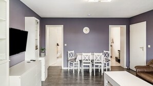 Una sala de estar moderna con un televisor de pantalla plana, una mesa de comedor con sillas, un sofá y un reloj en la pared.