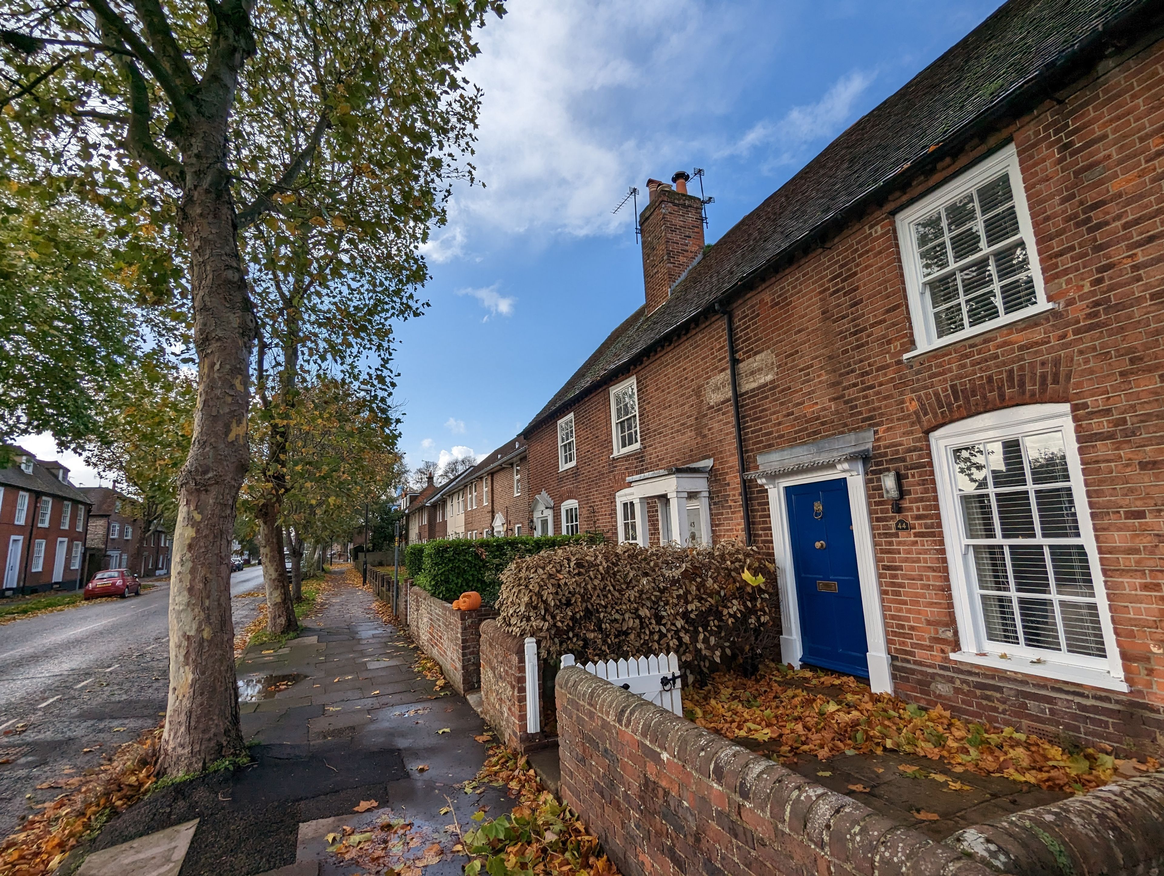 A row of traditional brick houses with a blue door, a white picket fence, and a tree-lined street.