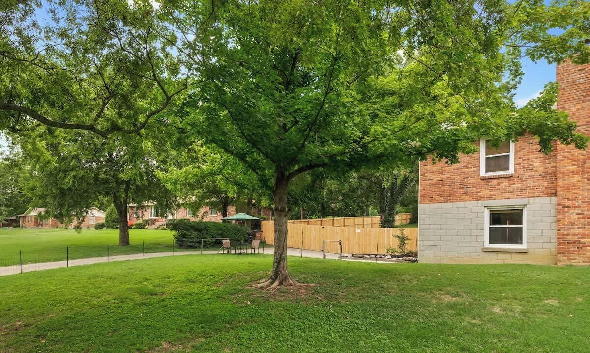 A tree-lined courtyard with a brick building, a wooden fence, and a grassy lawn.