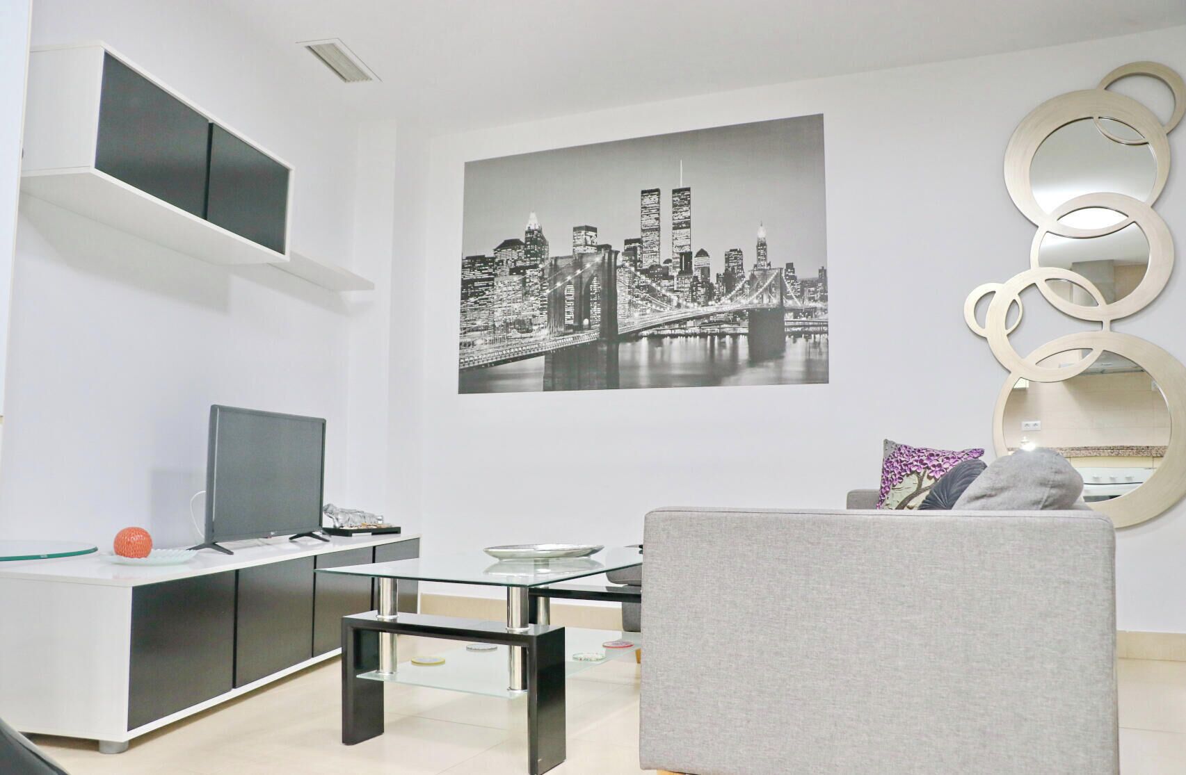 Una sala de estar moderna con un televisor de pantalla plana, una mesa de centro de vidrio, un sofá y una pintura de un paisaje urbano en la pared.