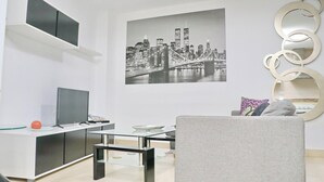 Una sala de estar moderna con un televisor de pantalla plana, una mesa de centro de vidrio, un sofá y una pintura de un paisaje urbano en la pared.