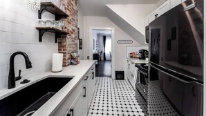 A modern kitchen with a brick accent wall, a coffee bar, and a refrigerator. - Roaring Riverside - Art Deco Luxe - 2 Kings! (Wichita)