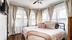 A bedroom with a bed, curtains, a ceiling fan, and a TV mounted on the wall. - Roaring Riverside - Art Deco Luxe - 2 Kings! (Wichita)
