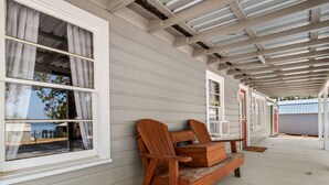 A covered porch with wooden chairs and a bench, white siding, and a view through the windows. - Pleasure Point Place WATERFRONT Cottage (Florien)
