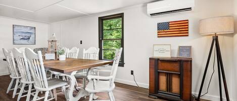 Un comedor con una mesa de madera y sillas blancas, un equipo de aire acondicionado, una lámpara y un ventanal con vistas a zonas verdes.