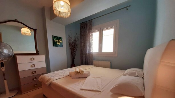 Ein Schlafzimmer mit Bett, Kommode, Spiegel, Fenster mit Vorhängen und einer Nachttischlampe.