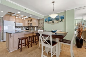 A modern kitchen with wooden cabinets, a central island with bar stools, a dining table, and a hanging light fixture. - Discover a top-tier beach getaway at SEA MARSH II 1003 (North Myrtle Beach)