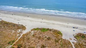 Ein Strand mit breitem Sandstrand, etwas Vegetation im Vordergrund und einem klaren, blauen Himmel.