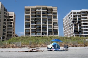 Hotel davant de la platja amb diversos balcons, una tenda i cadirees de platja.