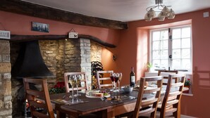 Un coin repas agrémenté d’une cheminée en pierre, de chaises en bois, d’une table dressée pour deux personnes et d’une fenêtre donnant sur les voitures garées.