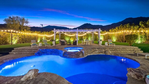 A swimming pool with a hot tub, surrounded by a pergola and string lights, set against a mountainous backdrop. - The Desert Castle with a Movie Theatre, golf, game room, sleeps 23 (Palm Desert)