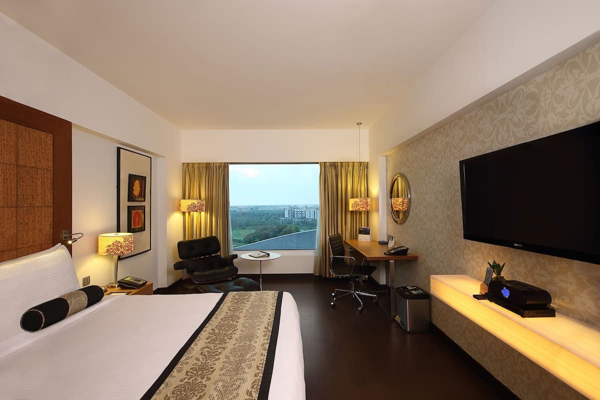 A hotel room with a bed, desk, chair, TV, and a view of a cityscape.