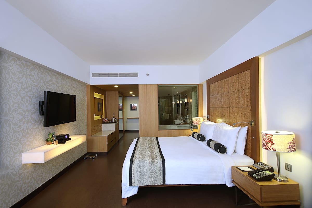 A modern hotel room with a large bed, a flat-screen TV, a bedside table with a lamp, and a bathroom visible through an open door.