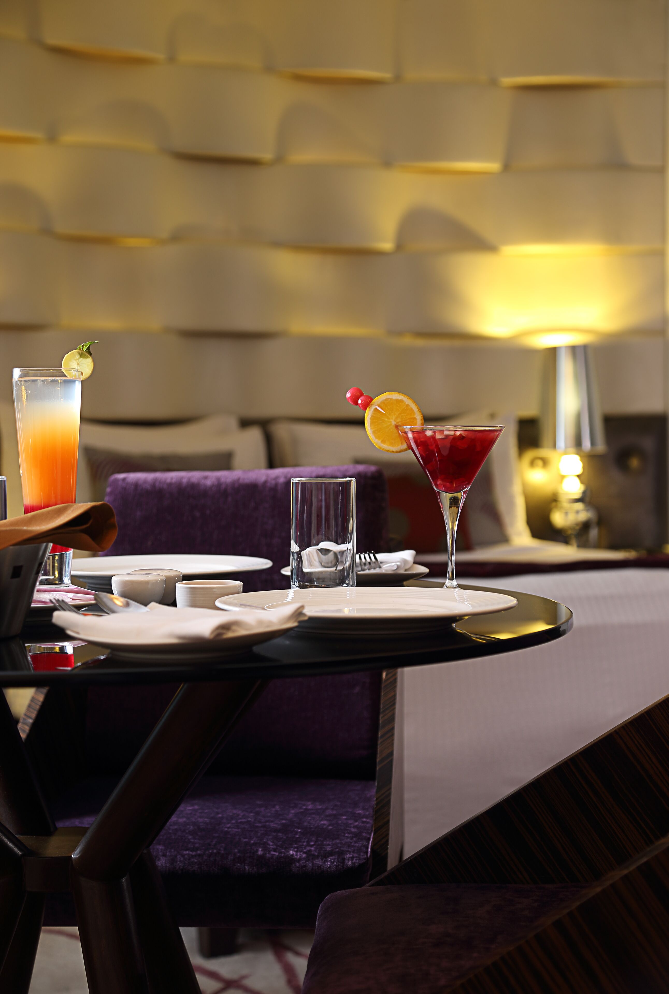 A dining table set with a martini, a glass of orange juice, and a water glass, with a purple upholstered chair and a lamp in the background.