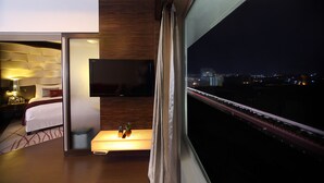 Una habitación de hotel moderna con una cama, un televisor montado en la pared, una mesita de noche con un control remoto y fruta, y vistas a un paisaje urbano nocturno.