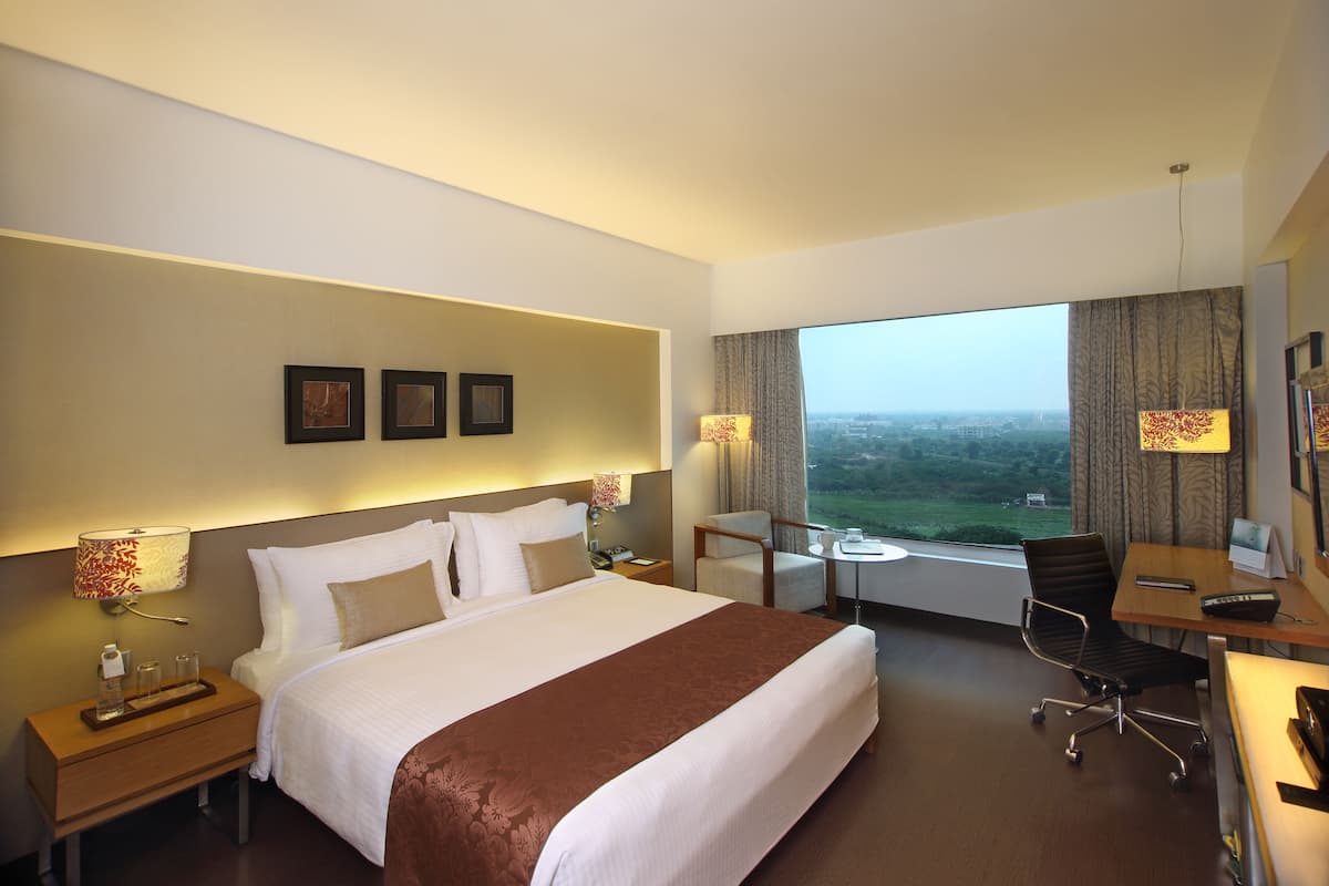 A hotel room with a large bed, a desk with a chair, a view of the city, and a window with curtains.
