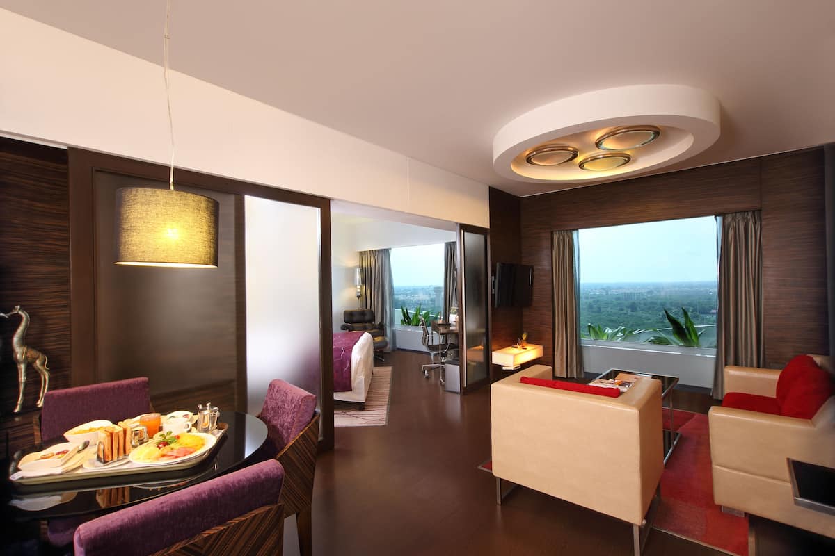 A modern hotel room with a dining area, a living area with a sofa and armchair, a bed, and a large window with a view.