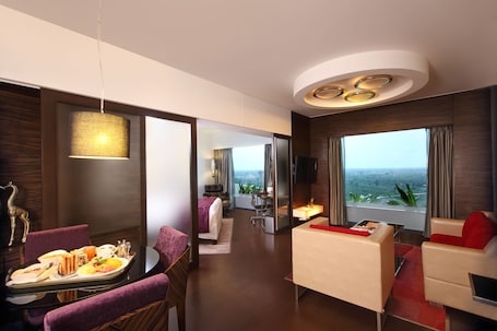 A modern hotel room with a dining area, a living area with a sofa and armchair, a bed, and a large window with a view.